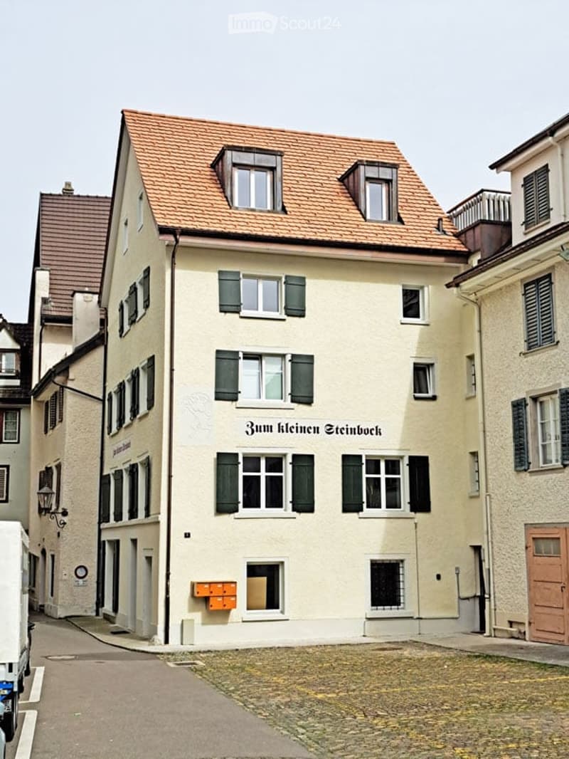 A three-story residential building with a red-tiled roof, multiple windows with green shutters, and a sign reading "Zum kleinen Steinbock".