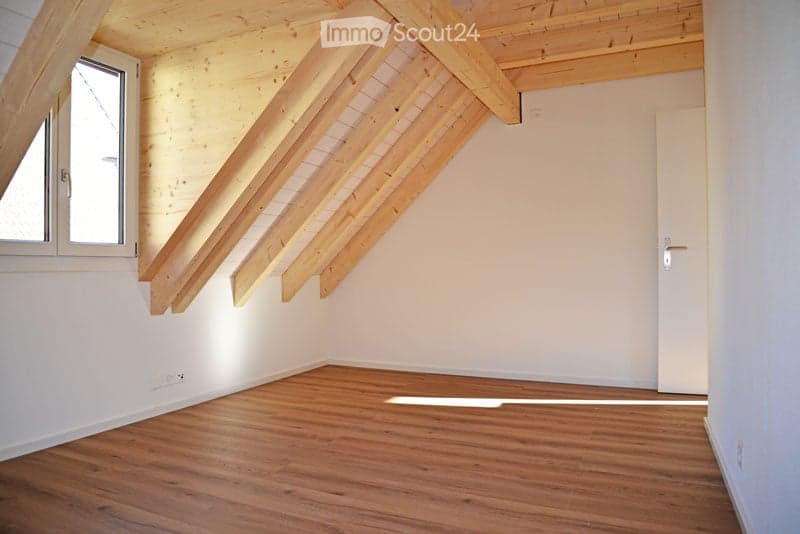 Empty attic room with wooden beams, wooden floor, white walls, and a window letting in natural light.