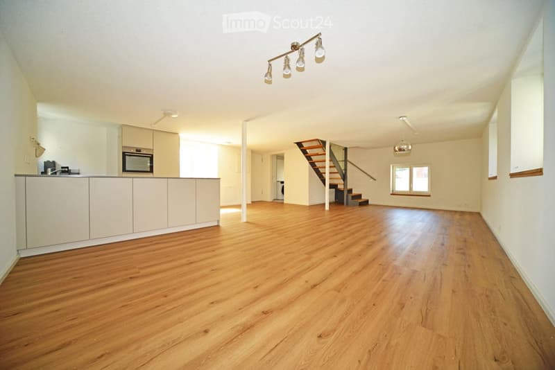 Open-plan unfurnished room with light wooden flooring, white walls, and a staircase leading to an upper level. Kitchen area with cabinets and appliances is visible on the left side.