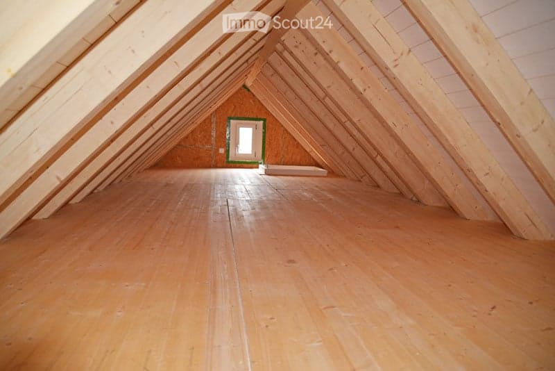 Empty attic space with wooden beams and flooring, featuring a small window at the far end.
