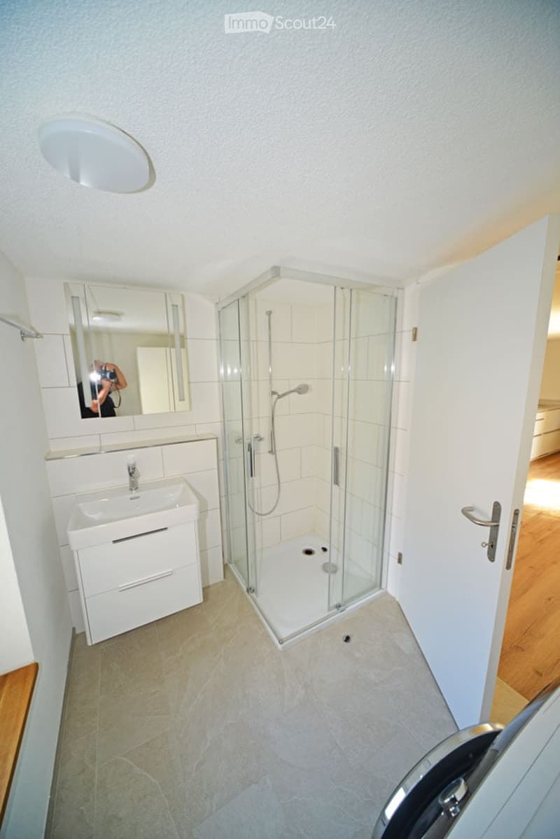 Modern bathroom with a white sink, mirror, and glass-enclosed shower stall. The space features light-colored tiles and a sleek design.