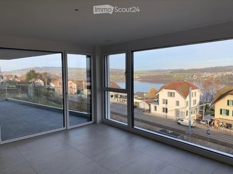 Bright, empty room with large floor-to-ceiling windows offering a scenic view of a lake and surrounding landscape. The space features a modern design with a tiled floor and a glass balcony door.