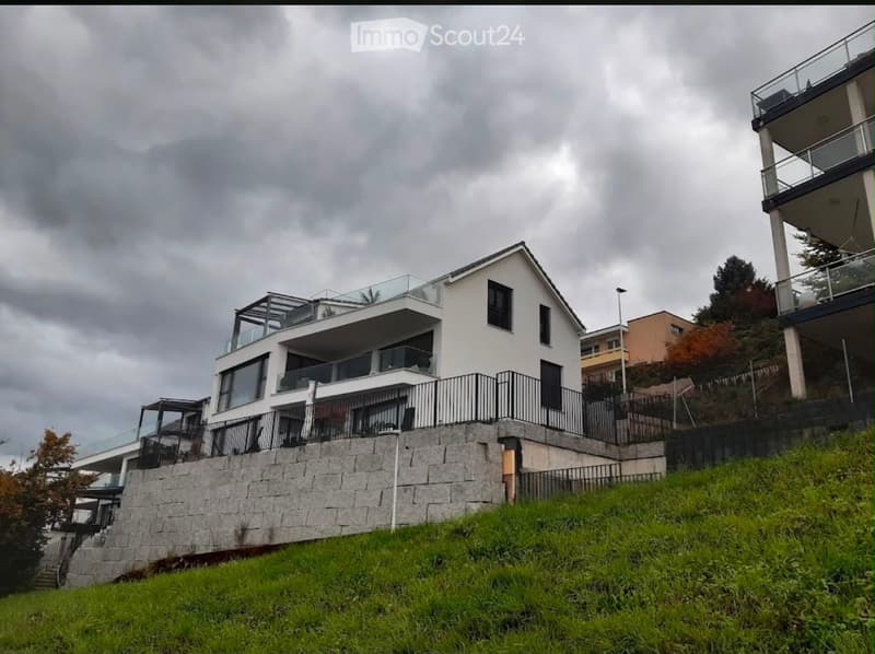 Modern two-story house with white exterior, large windows, and multiple balconies, situated on a raised ground floor with stone retaining wall and green lawn.