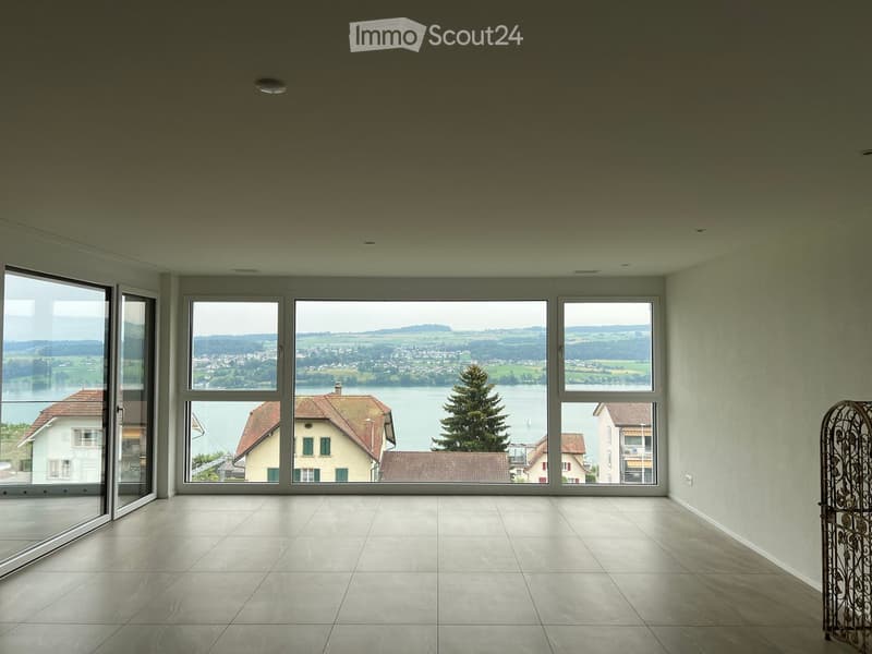 Empty room with large windows offering a scenic view of a lake and surrounding landscape. The room features tiled flooring and modern recessed lighting.