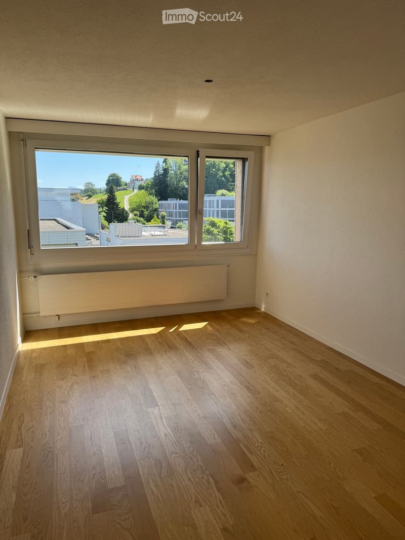 Empty room with wooden flooring, large windows, and a radiator under one window. The view shows greenery and buildings outside.