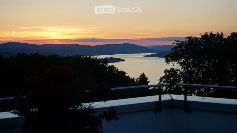 Balcony with a scenic view of a lake and distant mountains during sunset, surrounded by trees and a railing.