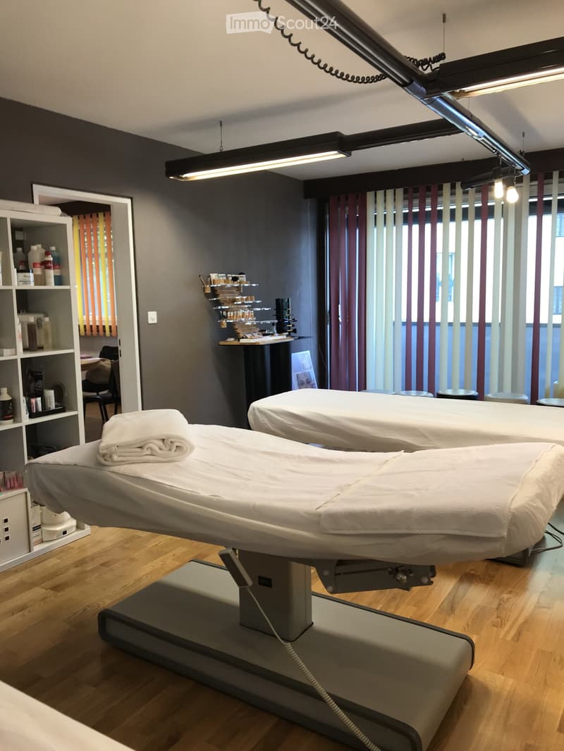 Modern spa treatment room with two treatment tables, shelves with bottles and products, a chair, and a mirror. Large windows with vertical blinds provide natural light.