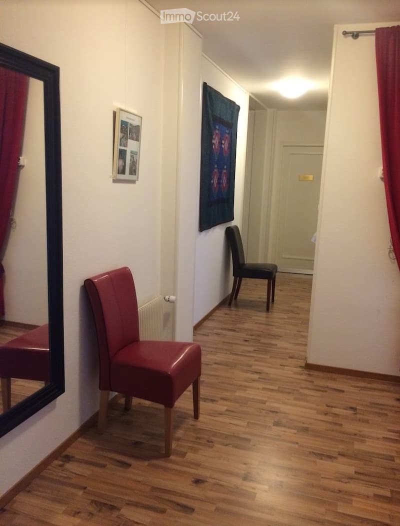 A narrow hallway with wooden flooring, two chairs, a mirror, and a picture frame on the wall.