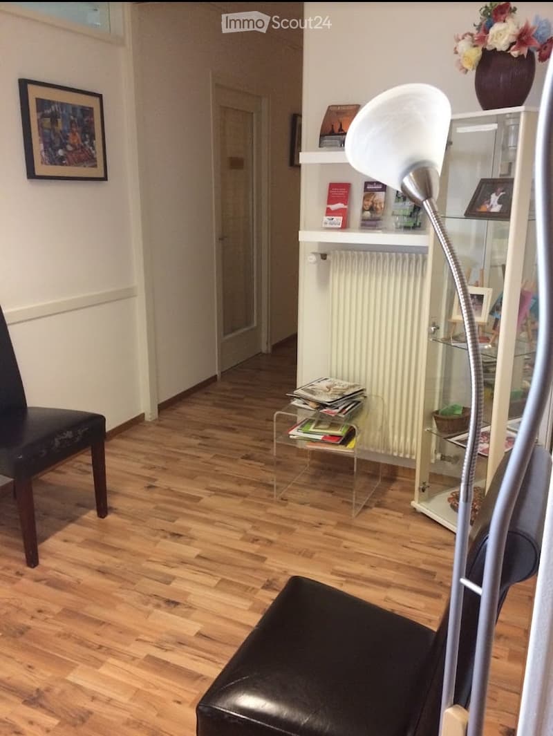 A modern living room with wooden flooring, a black leather chair, a glass coffee table with books, a white floor lamp, a white shelf with books and decorative items, and a vase with flowers on top.