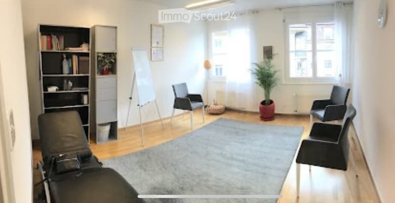 A modern, well-lit room with wooden flooring, multiple black chairs, a bookcase, a potted plant, and a whiteboard. The space is organized and appears to be used for meetings or collaborative work.