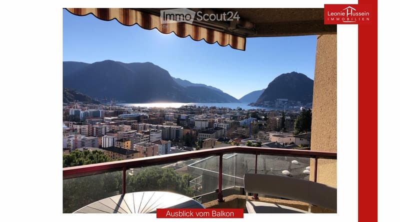 Balcony with a panoramic view of a cityscape, lake, and mountains under a clear blue sky.
