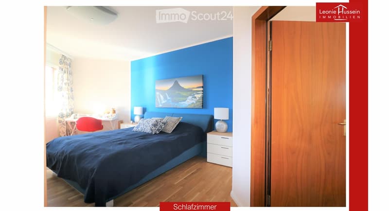 Bright bedroom with blue accent wall, wooden flooring, large bed with dark blue bedding, nightstand with lamp, desk with chair, and a scenic picture on the wall.