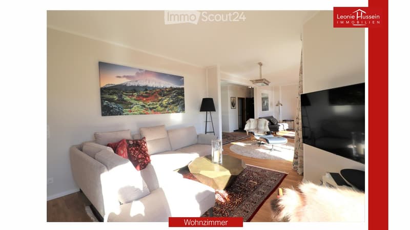 Bright living room with a large sectional sofa, coffee table, floor lamp, and television. The space features a large painting on the wall and a patterned rug on the wooden floor.