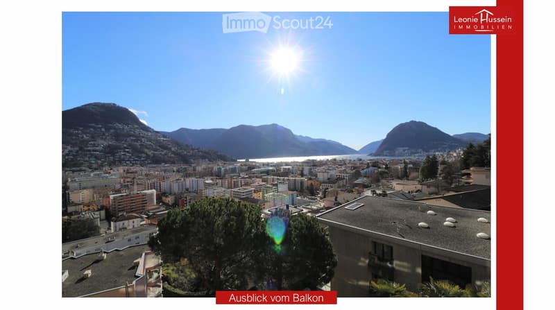 Panoramic view from a balcony showing a cityscape with buildings, a lake, and mountains under a clear blue sky.