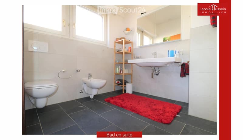 Modern bathroom with white tiles, dark grey floor, toilet, sink, mirror, wooden shelf, and red rug.