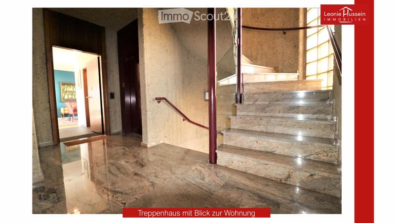 Spacious hallway with polished marble floors, a staircase with a polished metal railing, and an open doorway leading to a room with a red chair and a painting on the wall.