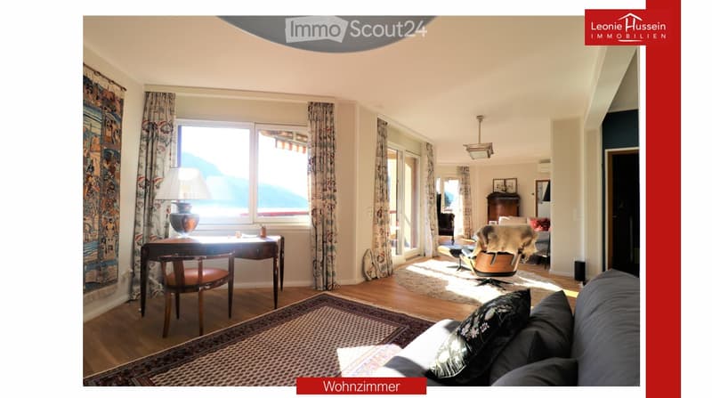 Bright living room with large windows offering a scenic view, furnished with a sofa, chair, table, lamp, and curtains.