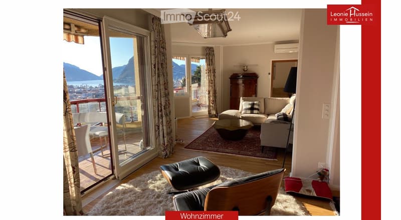 Bright living room with large windows and a view of the lake and mountains, featuring a sofa, coffee table, ottoman, chairs, and a dining area with a table and chairs.