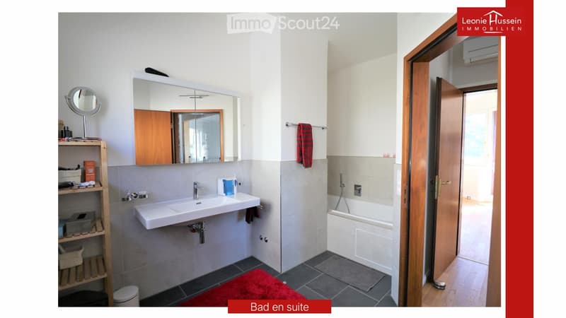 Modern bathroom with a white sink, large mirror, bathtub, and wooden shelf. Includes a handheld shower, towel, and a small red rug on the floor.