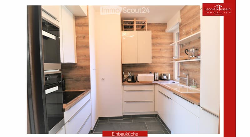 Modern kitchen with white cabinets, wooden accents, stainless steel appliances, and a built-in oven. Features a sink with a modern faucet, shelves, and various small appliances.
