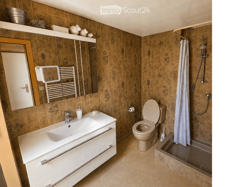 Bathroom with beige tiles, a white sink with a mirror, a toilet, a bathtub with a showerhead, and a towel rack. The walls have a floral pattern, and there is a shelf above the sink holding decorative items.
