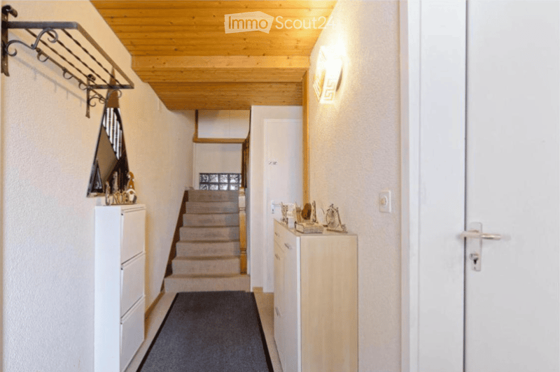 A narrow hallway with wooden ceiling, carpeted floor, and white walls. Features a staircase leading upwards, a coat rack with a coat hanging, and small decorative items on a cabinet.