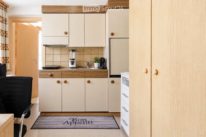 Modern kitchen with light-colored cabinets, a refrigerator, an oven, a blender, a coffee maker, and a sink. A small potted plant is placed on the counter, and a chair is visible in the foreground.