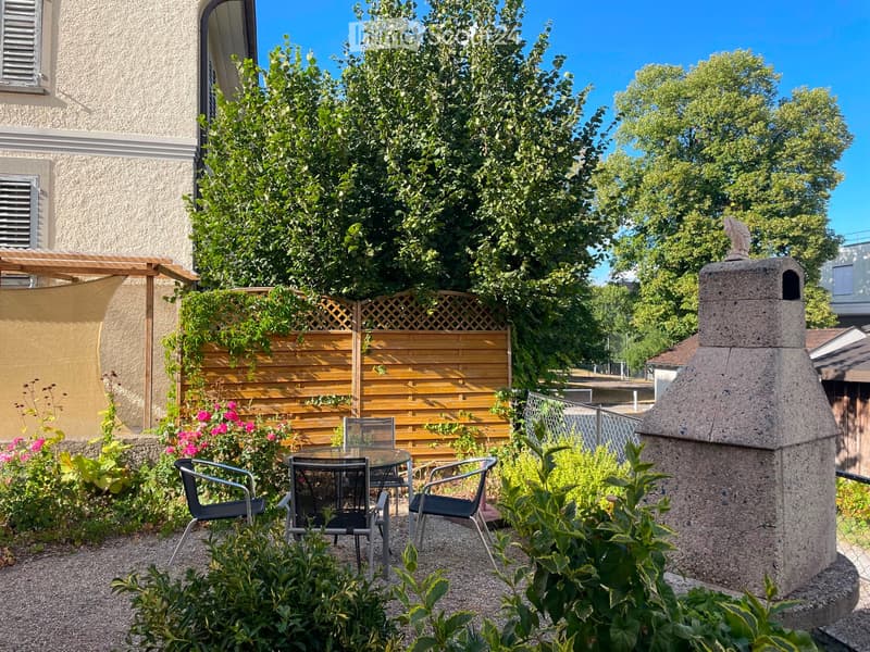 Garden area with a stone barbecue, a wooden fence, a pergola, and a small seating area with a table and chairs.