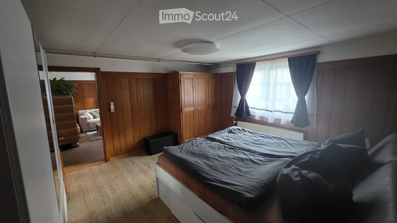 Cozy bedroom with wooden walls, a large bed with dark bedding, a wardrobe, a small living area with a sofa and table, and a plant near the doorway.