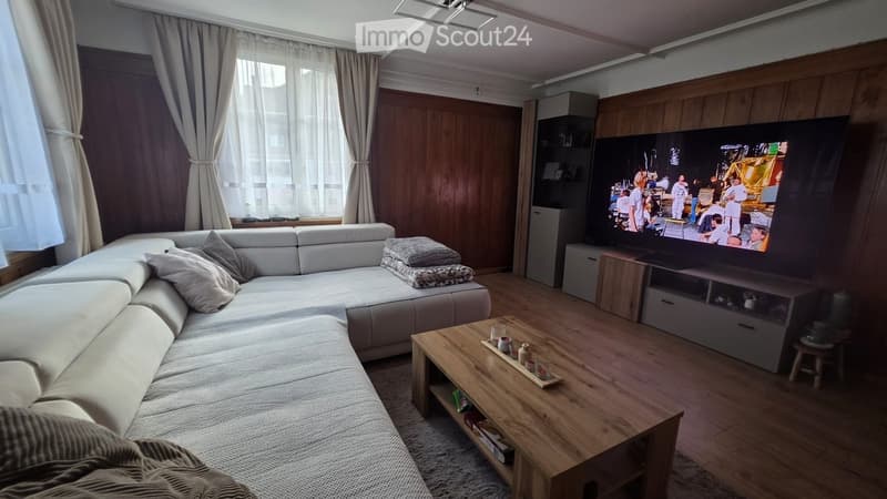 Modern living room with a large sectional sofa, wooden coffee table, flat-screen TV, and built-in cabinetry. Natural light from large windows with curtains.