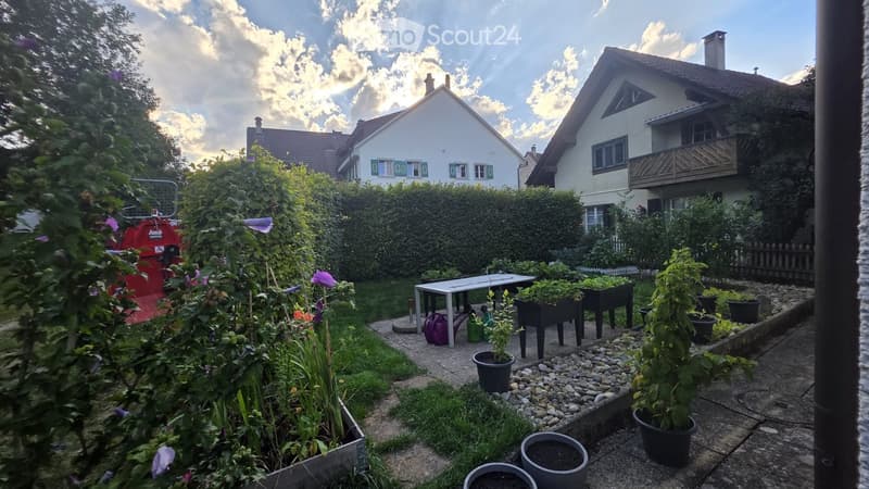 A well-maintained garden with potted plants, a small table, and a neatly trimmed hedge, surrounded by residential houses.