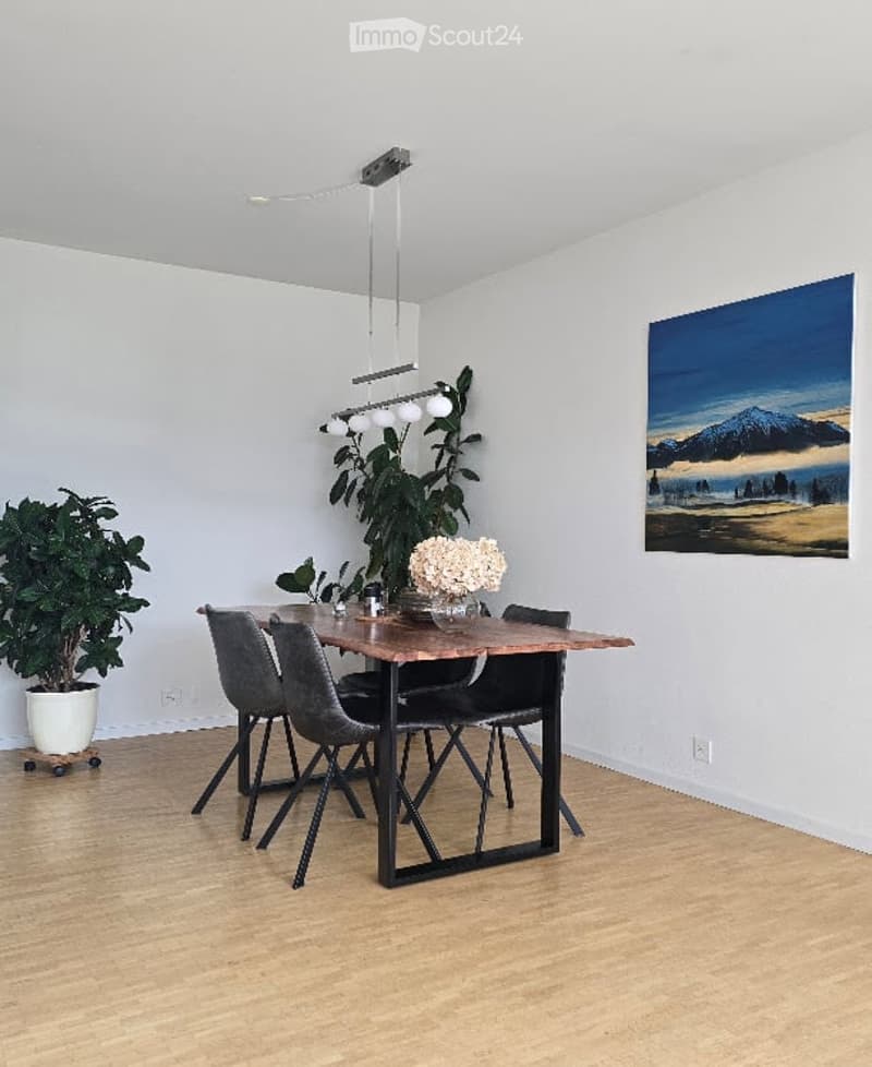 Modern dining area with a wooden table, four grey chairs, a hanging light fixture, and potted plants. A large painting of a landscape adorns the wall.