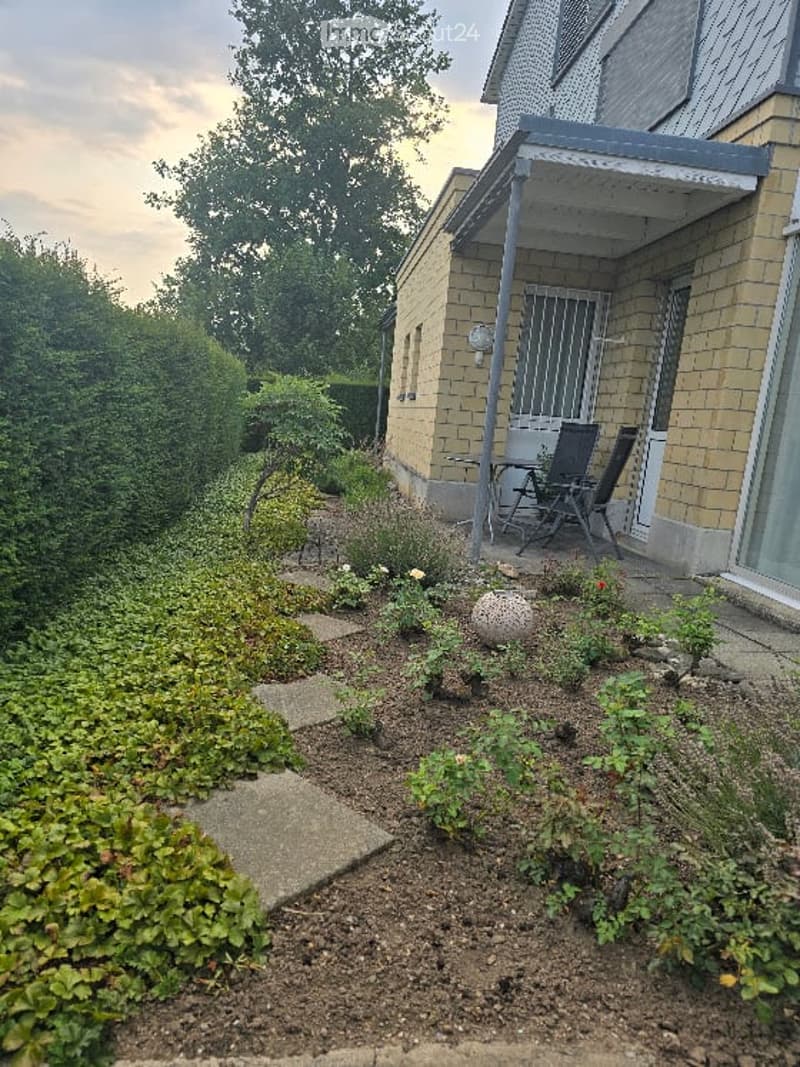 Well-maintained garden area with neatly trimmed hedges, stepping stones, and small flower beds. A patio with outdoor chairs is visible near the house entrance.