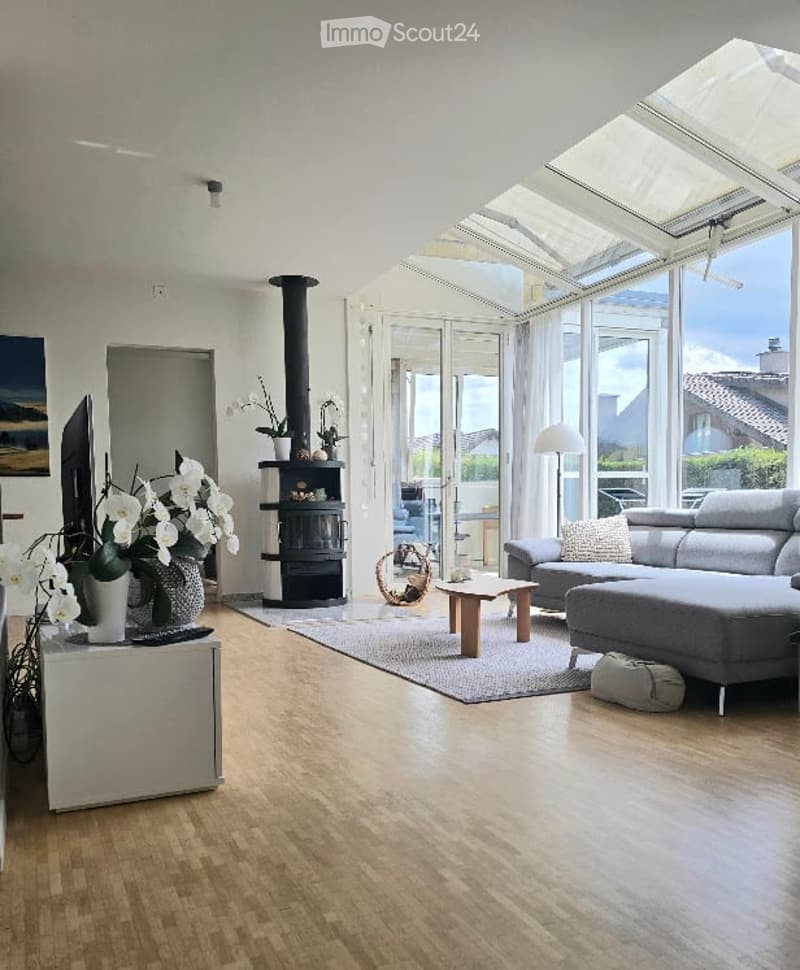Bright living room with wooden flooring, large windows, a fireplace, a grey sectional sofa, coffee table, floor lamp, and a television on a stand.