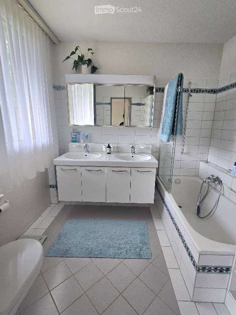 Modern bathroom with double sink, large mirror, bathtub, and a small plant on the windowsill.