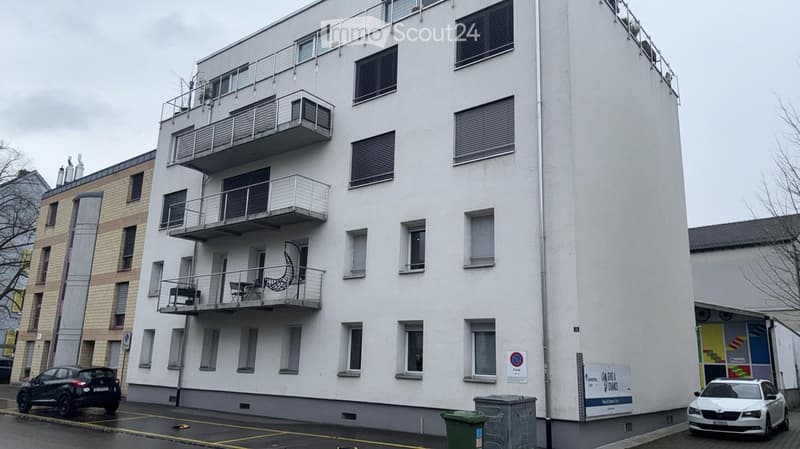 Modern multi-story apartment building with white facade, multiple balconies, and several windows. Balconies feature metal railings and some outdoor furniture. The building is situated in an urban area with parked cars and a trash bin visible in the foregr