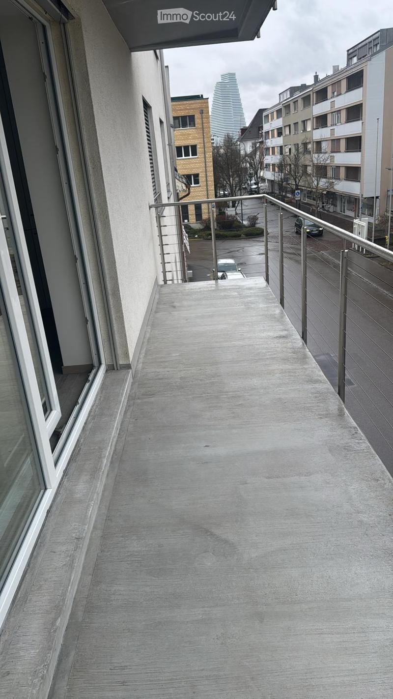 Balcony with metal railing, concrete floor, overlooking a city street with buildings and parked cars.