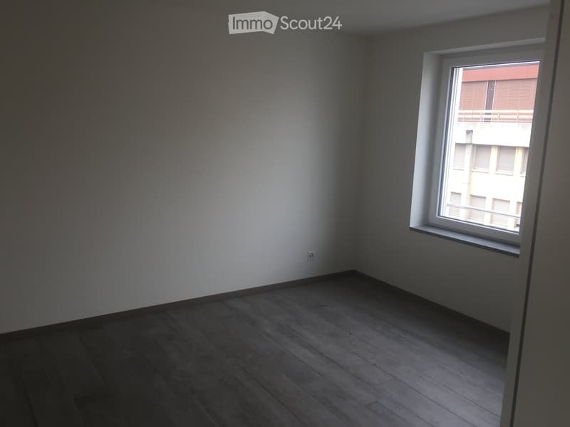 Empty room with wooden flooring, white walls, and a single window showing a view of a neighboring building.