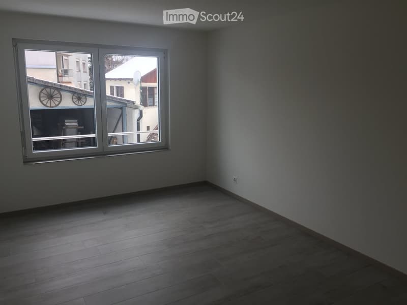 Empty room with wooden flooring and large windows, showing a view of the outside area.