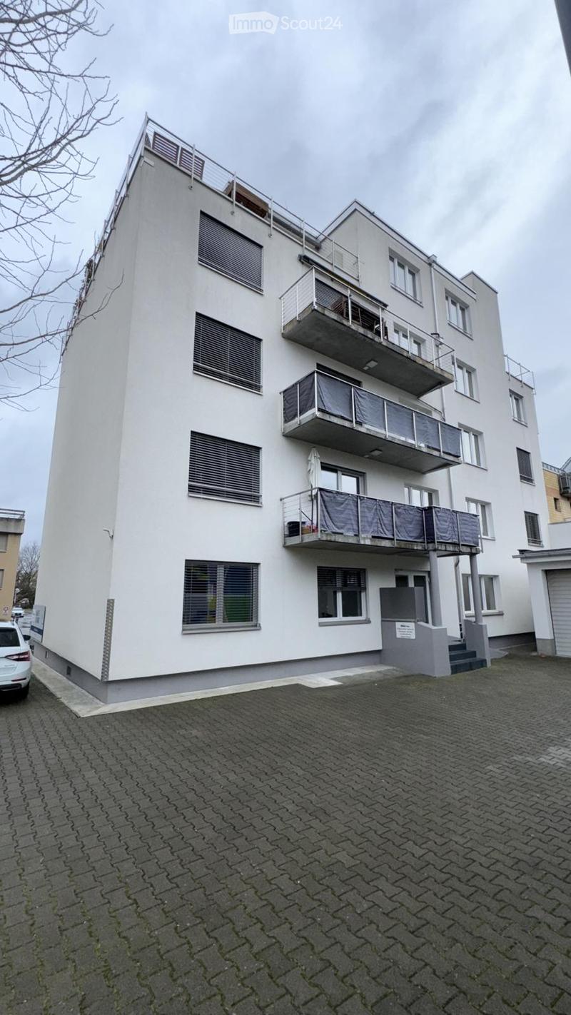 Modern multi-story apartment building with white facade, multiple balconies, and large windows. The building has a sleek, contemporary design with a paved parking area in front.