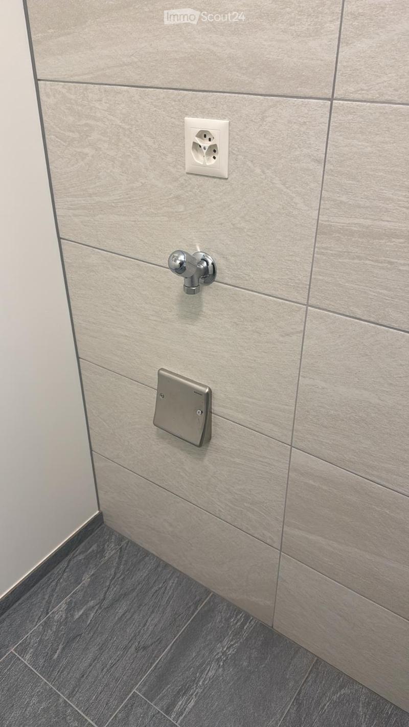 Walls covered with large rectangular tiles, floor covered with smaller rectangular tiles, electric outlet and pipe fitting on the wall