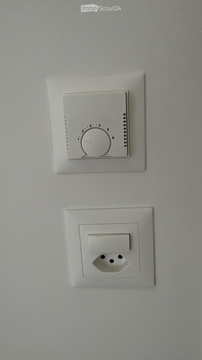 A plain wall with a thermostat and a power outlet, no furniture or furnishings present.