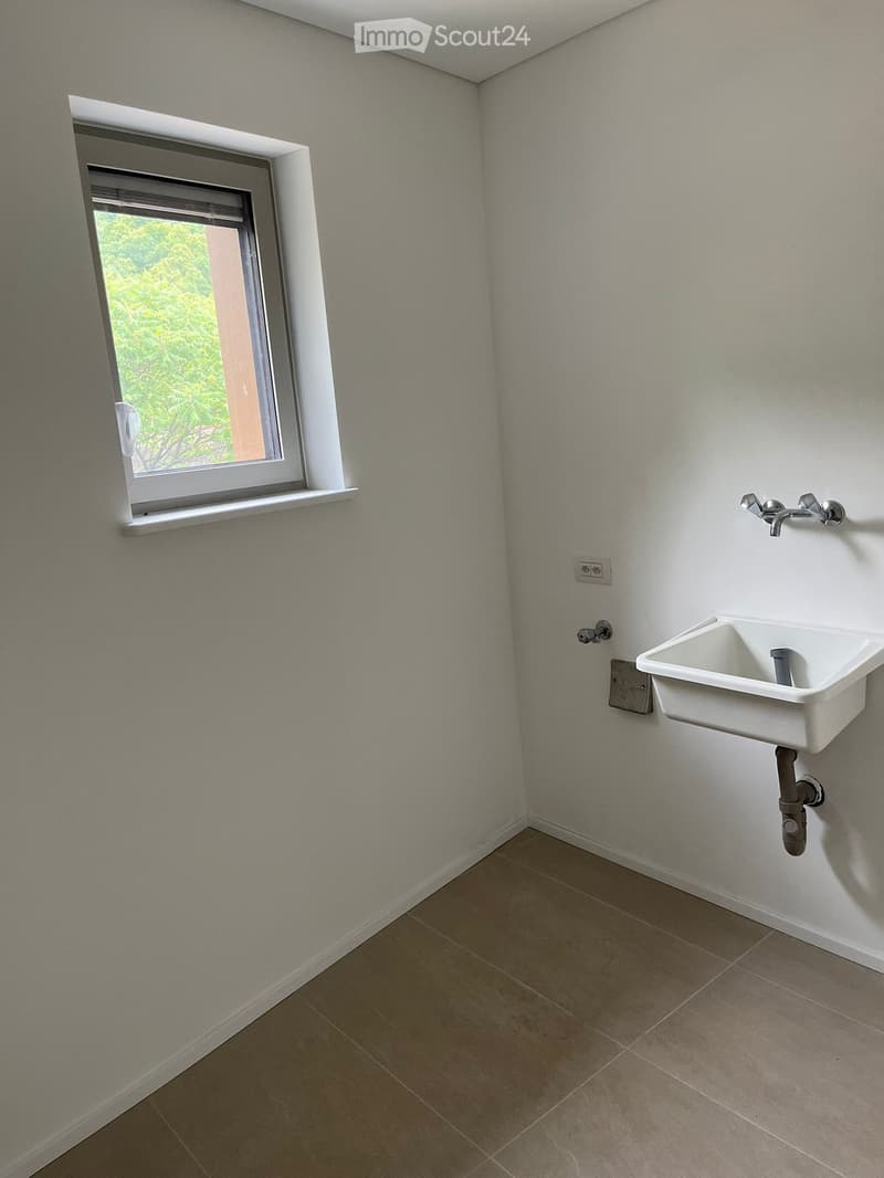 Empty bathroom with a white sink mounted on the wall, a window with a view of greenery, and beige tiled flooring.
