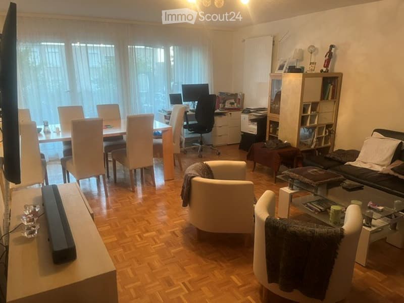 A well-lit living room with a mix of seating and a workspace. Features include a sofa, coffee table, several chairs, a dining table, a laptop on a desk, and a bookshelf with various items.