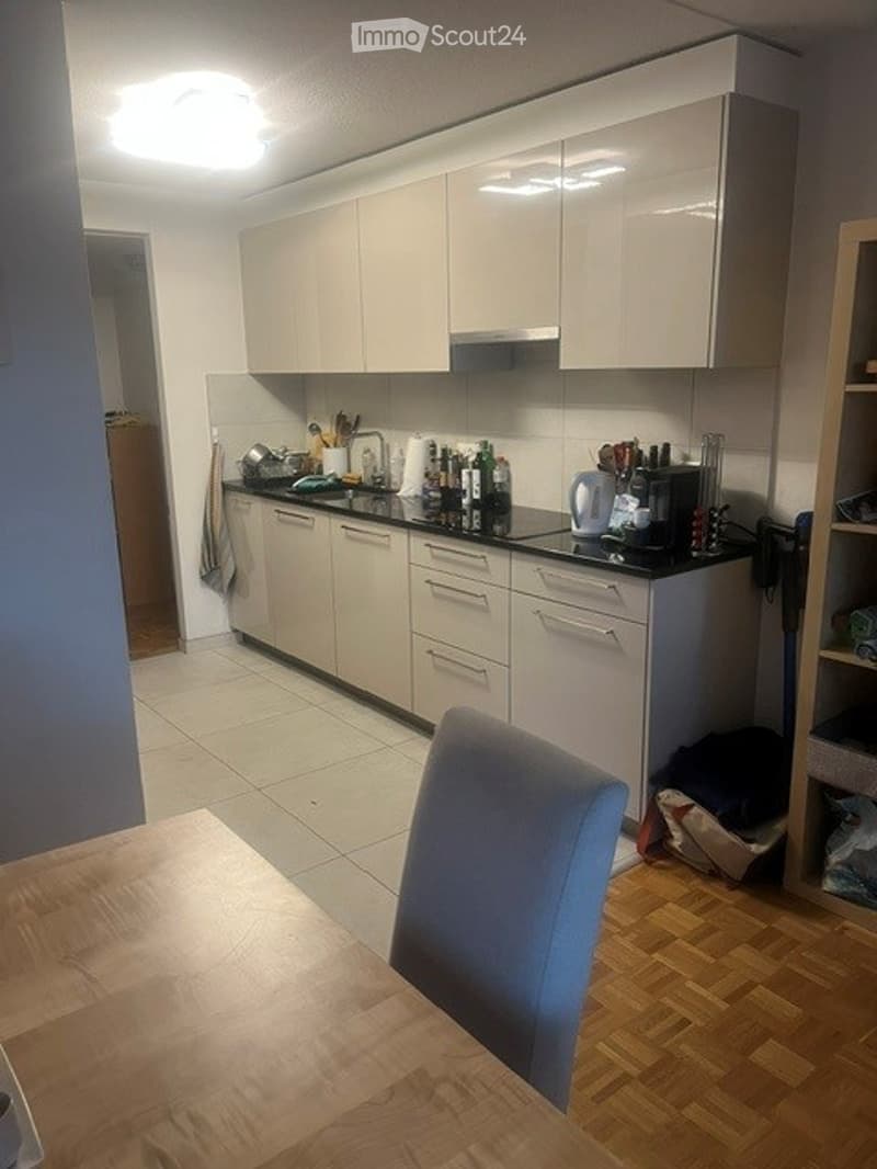 Modern kitchen with white cabinets, black countertops, and various appliances including a dishwasher, refrigerator, and microwave. A wooden dining table with a blue chair is visible in the foreground.