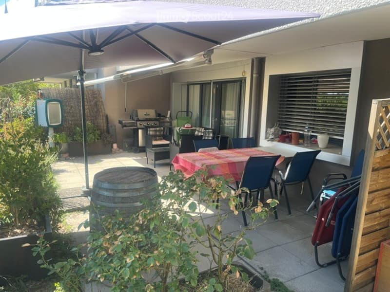 Outdoor patio area with a large umbrella, dining table with chairs, barbecue, and surrounding greenery.