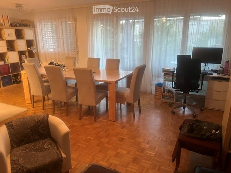 A well-lit dining and office area with a wooden dining table surrounded by chairs, a sofa, an office chair, a desk with a screen, and a small cupboard.