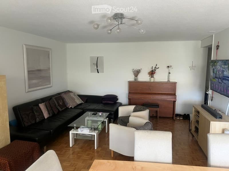 Living room with a black sectional sofa, glass coffee table, white chairs, piano, and a TV screen on a stand.