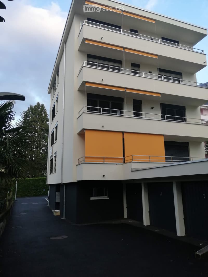 Modern multi-story building with white facade, orange awnings, and balconies. Features include multiple levels, railings, and a covered entrance area.