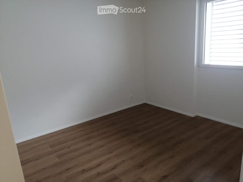 Empty room with light-colored walls and wooden flooring, featuring a single window with blinds on the right side.